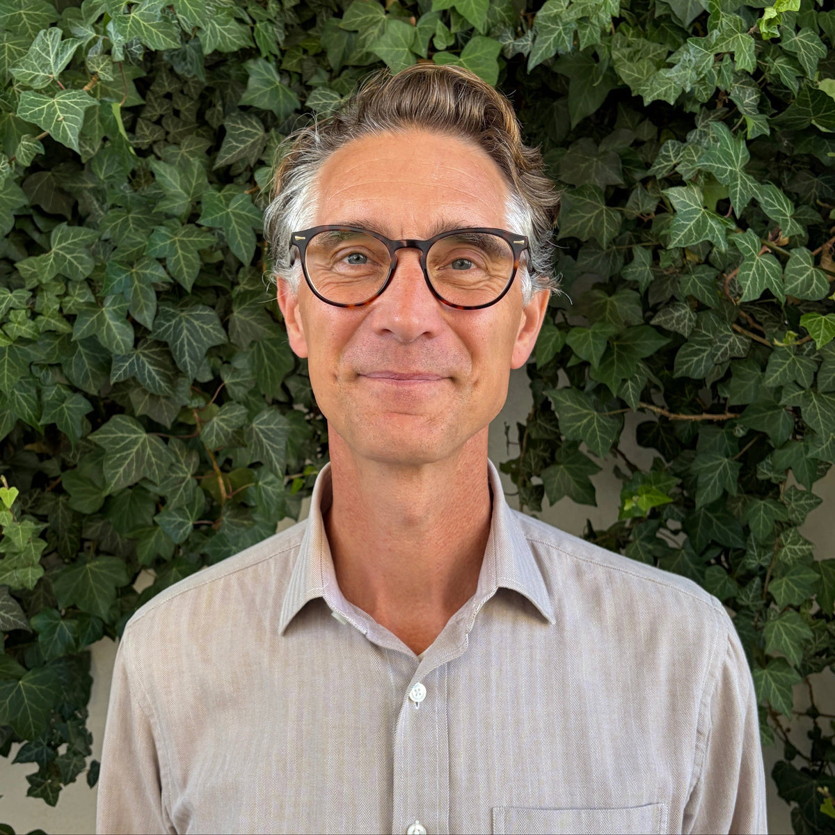 A man with glasses and styled hair stands in front of a dense green ivy wall, wearing a light beige button-down shirt.