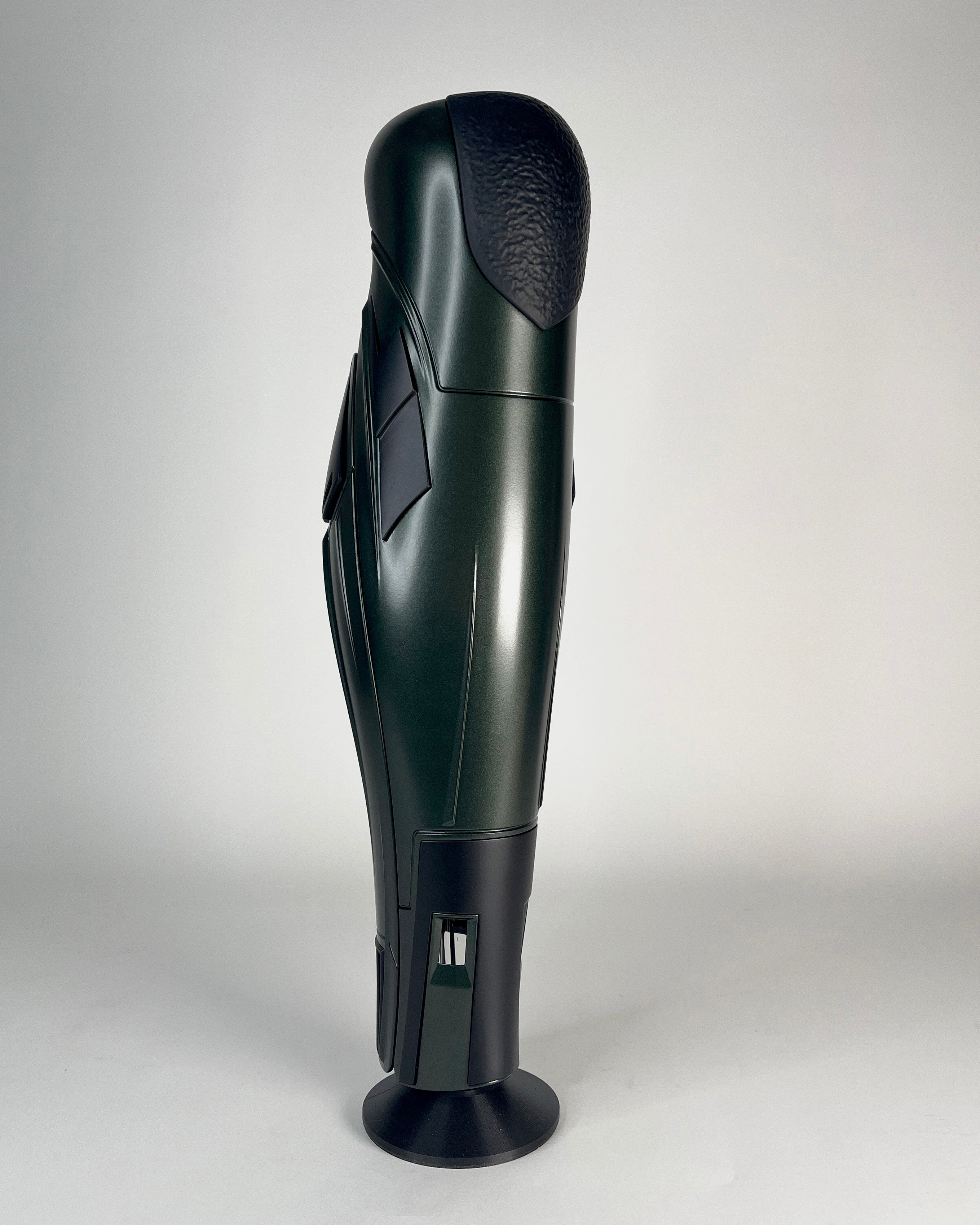 A dark green and black futuristic prosthetic leg or robotic limb standing upright on a circular base against a light gray studio background.