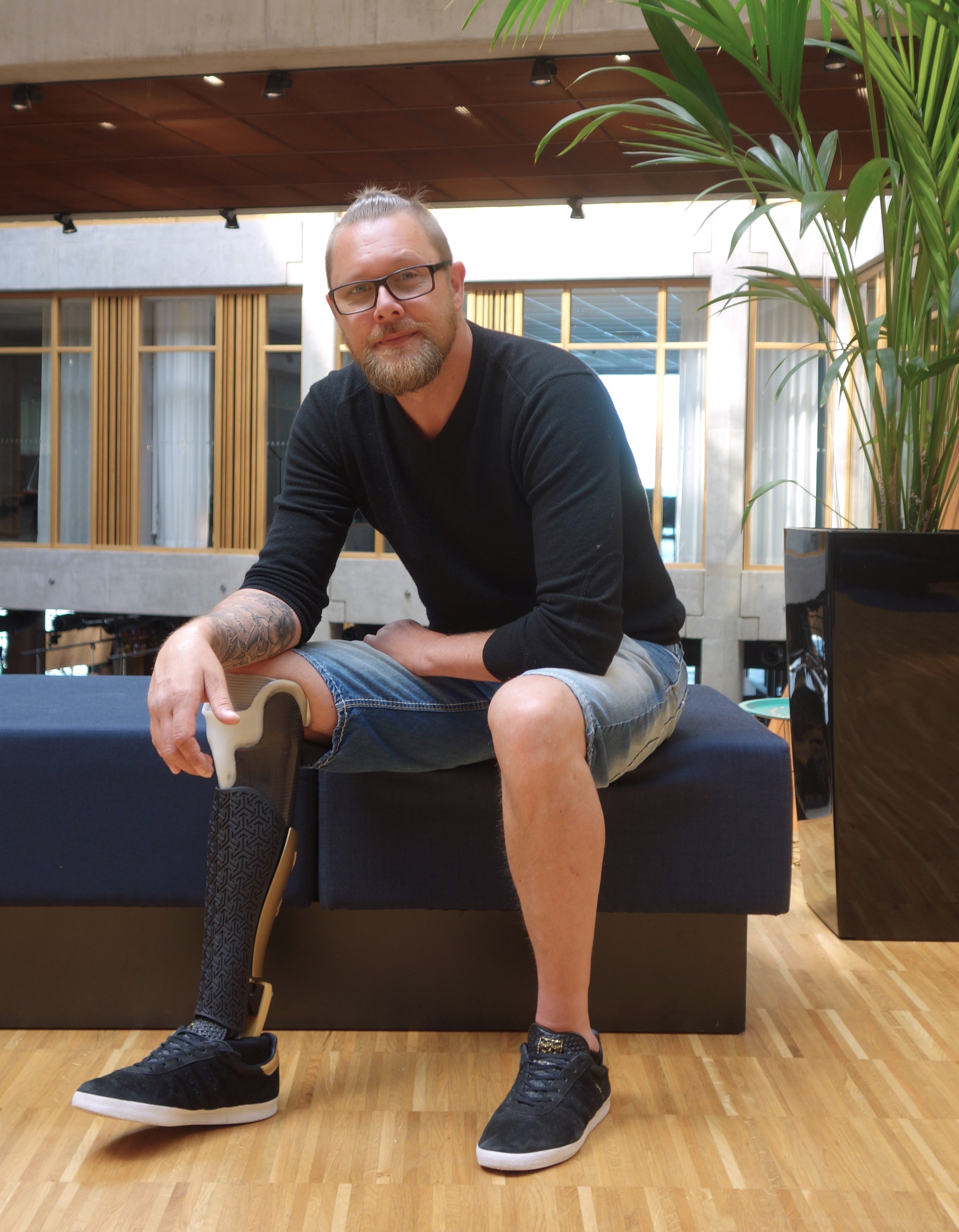 Man with a prosthetic leg sitting on a bench in an indoor setting with plants and windows.
