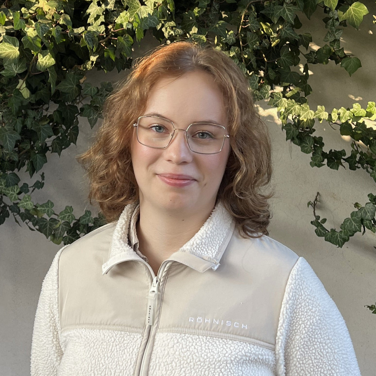 A woman with light brown hair and glasses wearing a beige and off-white fleece zip-up jacket, standing outdoors against a wall with green ivy.