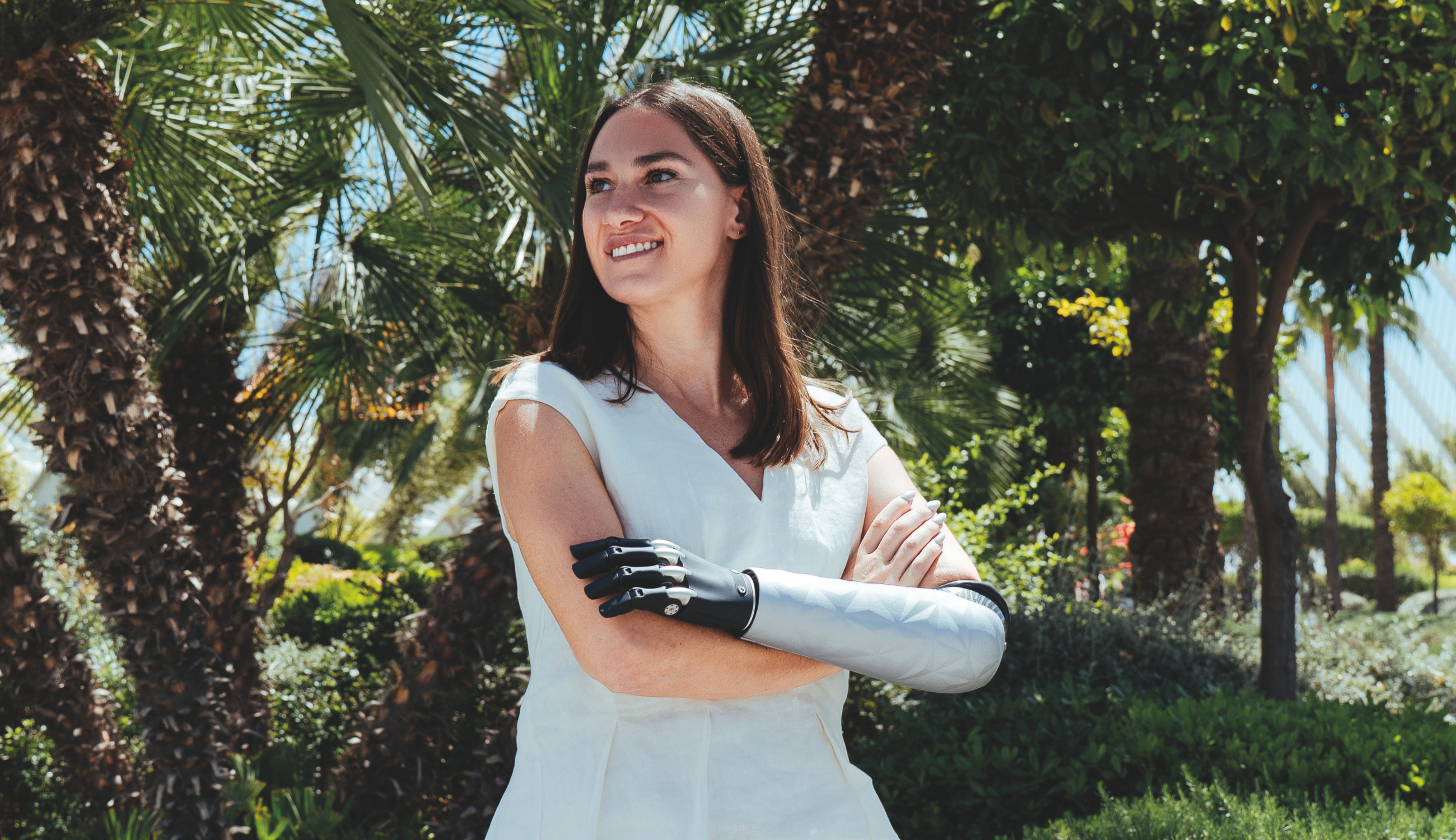A woman standing outdoors in a sunny garden with palm trees, wearing a white sleeveless dress and a silver prosthetic cover over a black arm prosthesis.