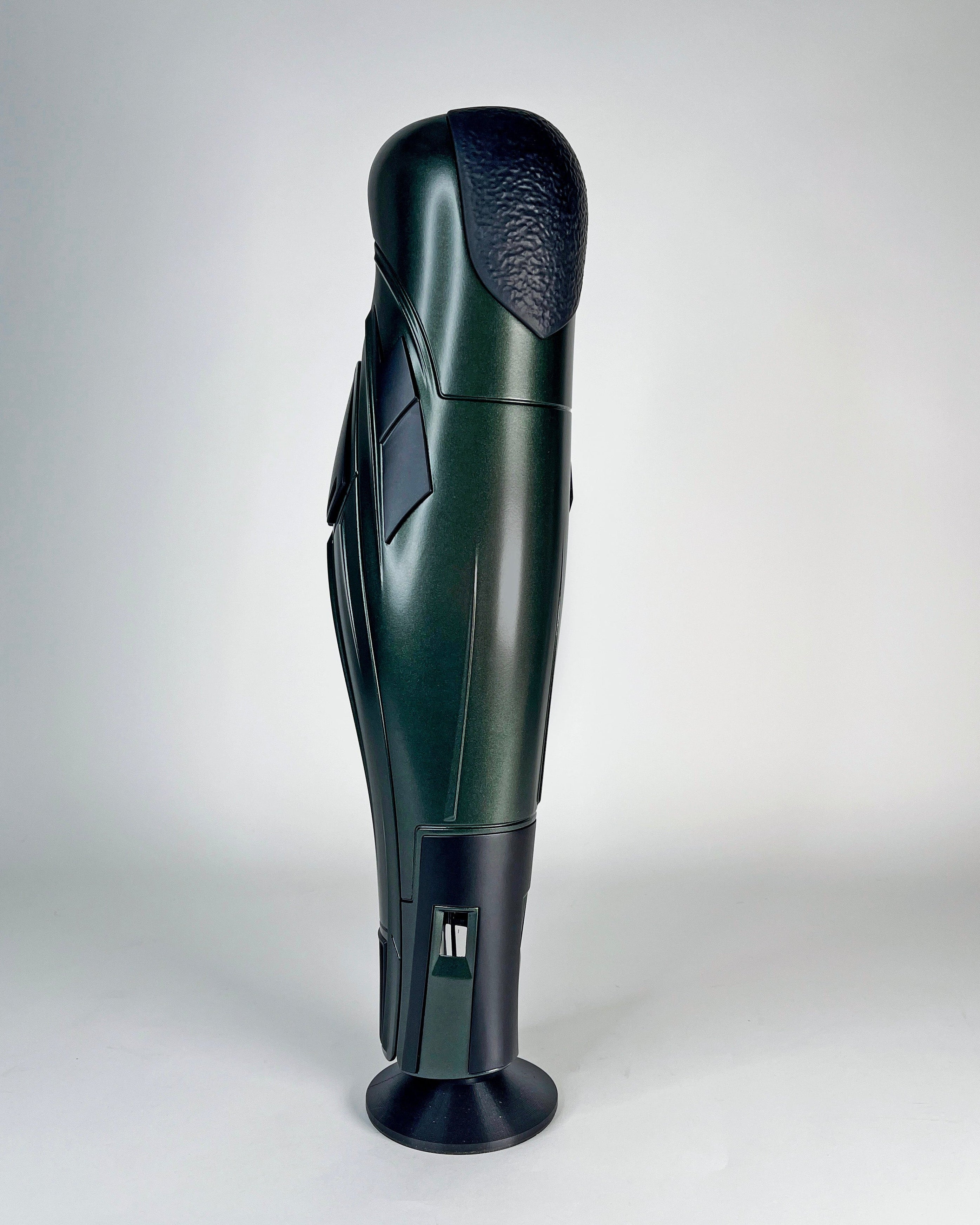A dark green and black futuristic prosthetic leg standing upright on a circular base against a light gray studio background.