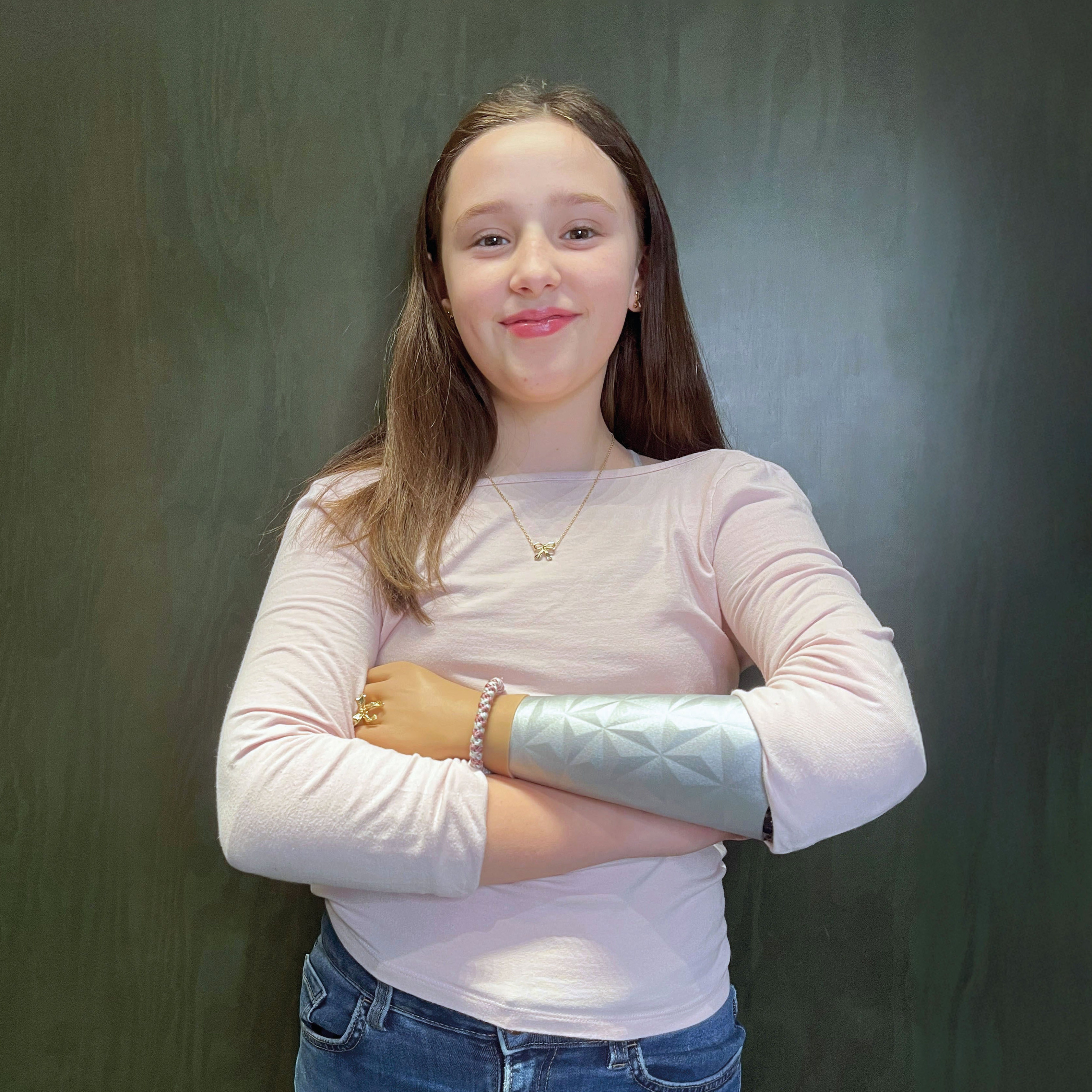 Young girl wearing a pink long-sleeve shirt, blue jeans and a silver coloured arm prosthesis against a dark background