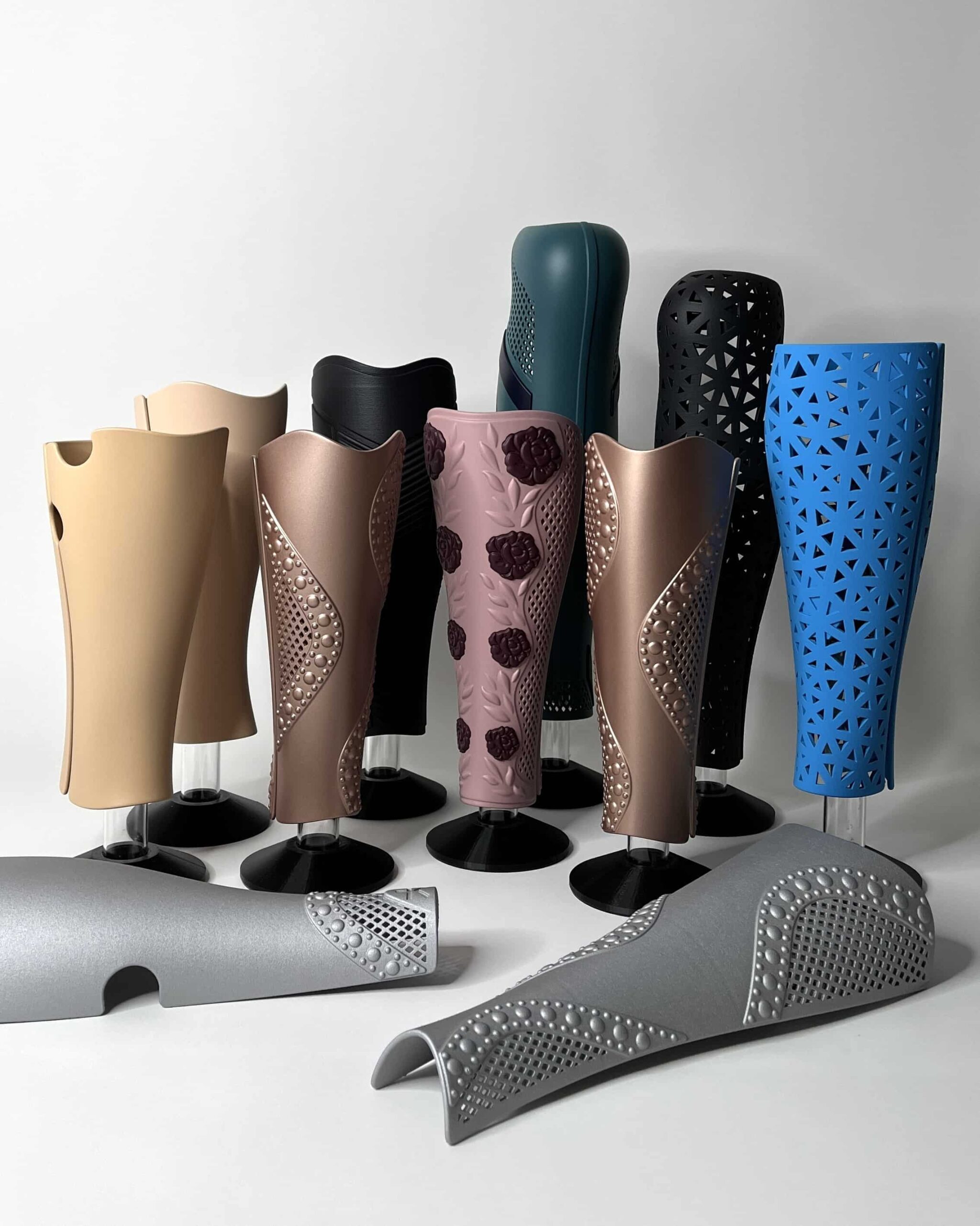 A collection of designed prosthetic covers displayed on black stands. The covers feature various colors and textures, including skin tones, metallic shades, blue, and green. The designs range from geometric patterns and perforated surfaces to floral decorations and pearl-like details. Two covers are placed in the foreground, while the others stand upright in the background against a white wall.
