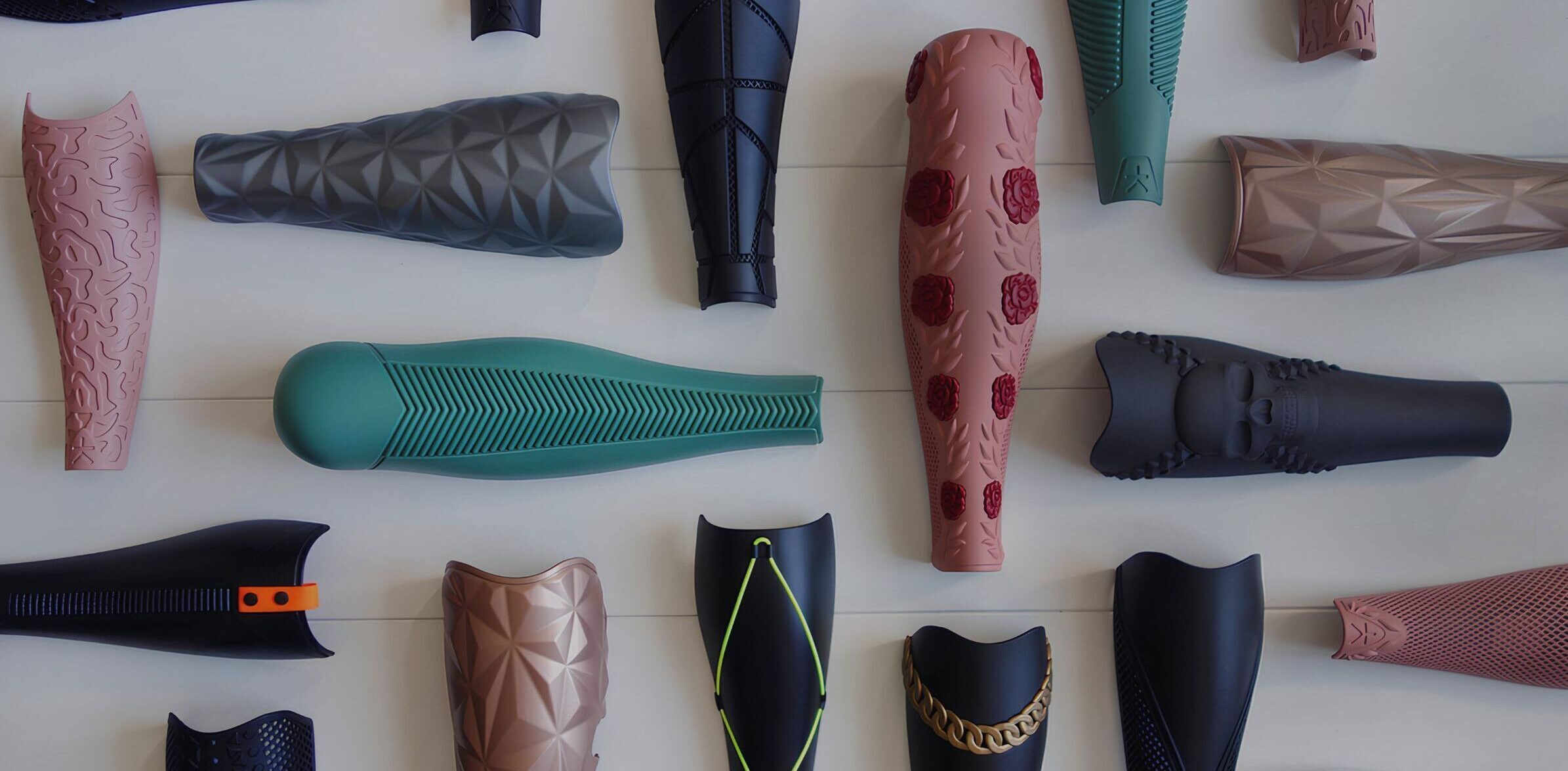 A collection of prosthetic leg covers displayed on a light-colored background. The covers feature a variety of intricate designs, textures, and colors, including geometric patterns, floral decor, chains, and skull motifs. The colors range from muted tones like black and gray to vibrant hues like green and pink.