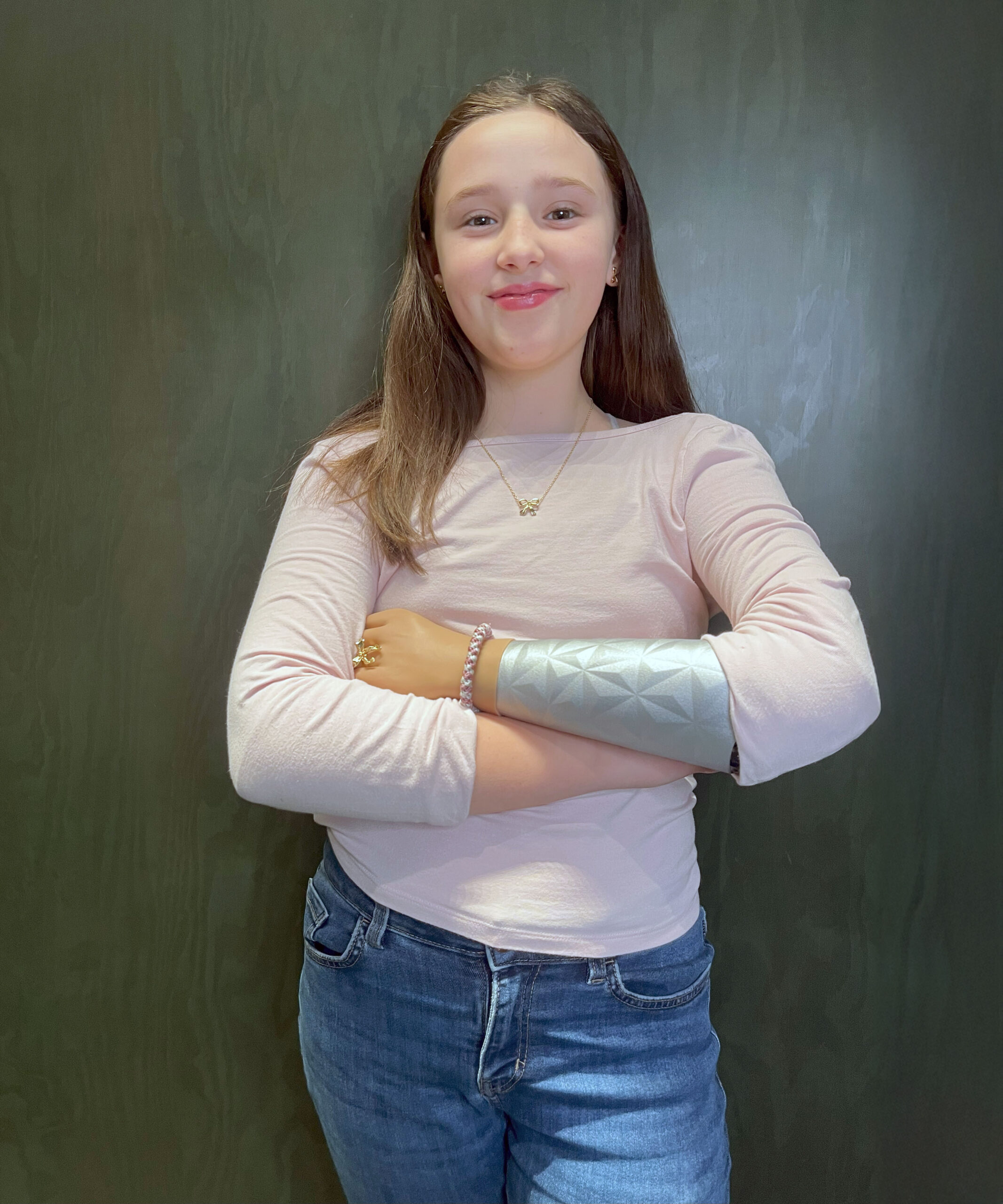 A young girl with long brown hair stands confidently with her arms crossed, smiling at the camera. She is wearing a light pink long-sleeve shirt, blue jeans, and a silver prosthetic arm cover with a geometric pattern. The background is a dark green wall.